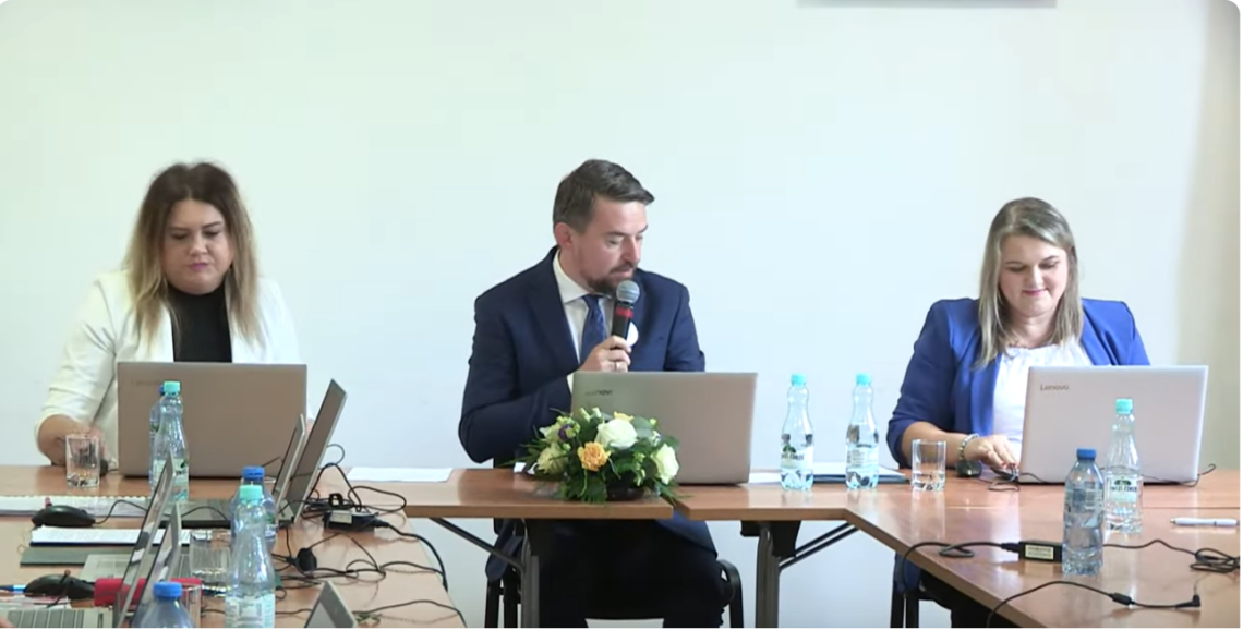 mężczyzna i po bokach siędzą dwie kobiety. Na stolikach stoją laptopy.
