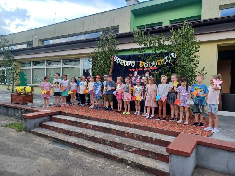 Dzieci stoją w szeregu przy schodach przedszkolnego tarasu, na nadgarstkach mają kolorowe motyle. W tle dekoracja z napisem: „Festyn Rodzinny”.