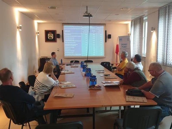 kilka osób siedzi przy stoe. Na ekranie wyświetlany jest slajd