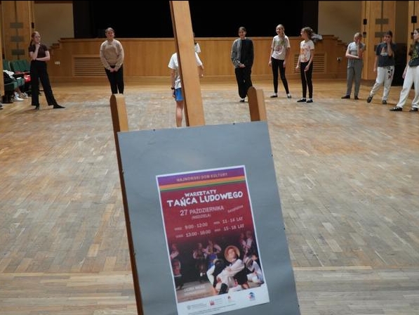 na pierwszym planie plakat promujący wydarzenie. W tle widać młodzież w sali kolumnowej domu kultury