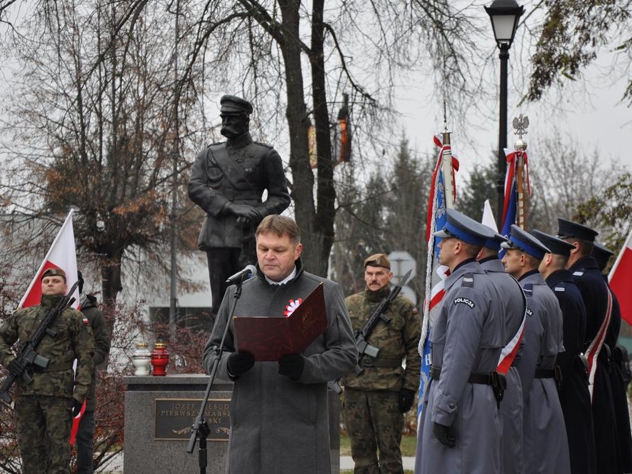 Mężczyzna stoi przy mikrofonie. Obok stoją poczty sztandarowe, w tle widać pominik Marszałka JózefaPiłsudskiego