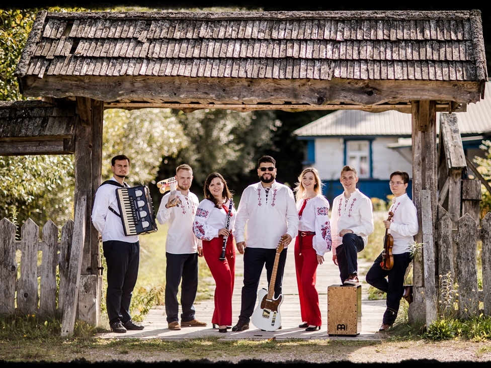 członkowie zespołu ubrani w ludowe stroje stoją w drewnianej bramie i trzymaja instrumenty. W tle widać chatę.