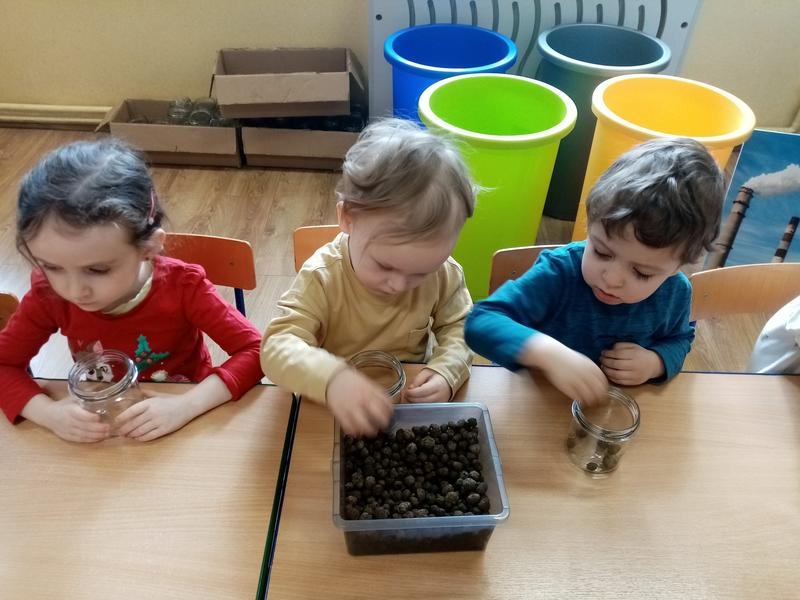 troje dzieci siedzi przy stoliku i wkłada do słoików produkty