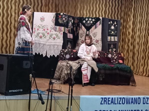 dwie osoby w stojach ludowych na scenie. W tle widać zawieszone haftowane makaty i chusty