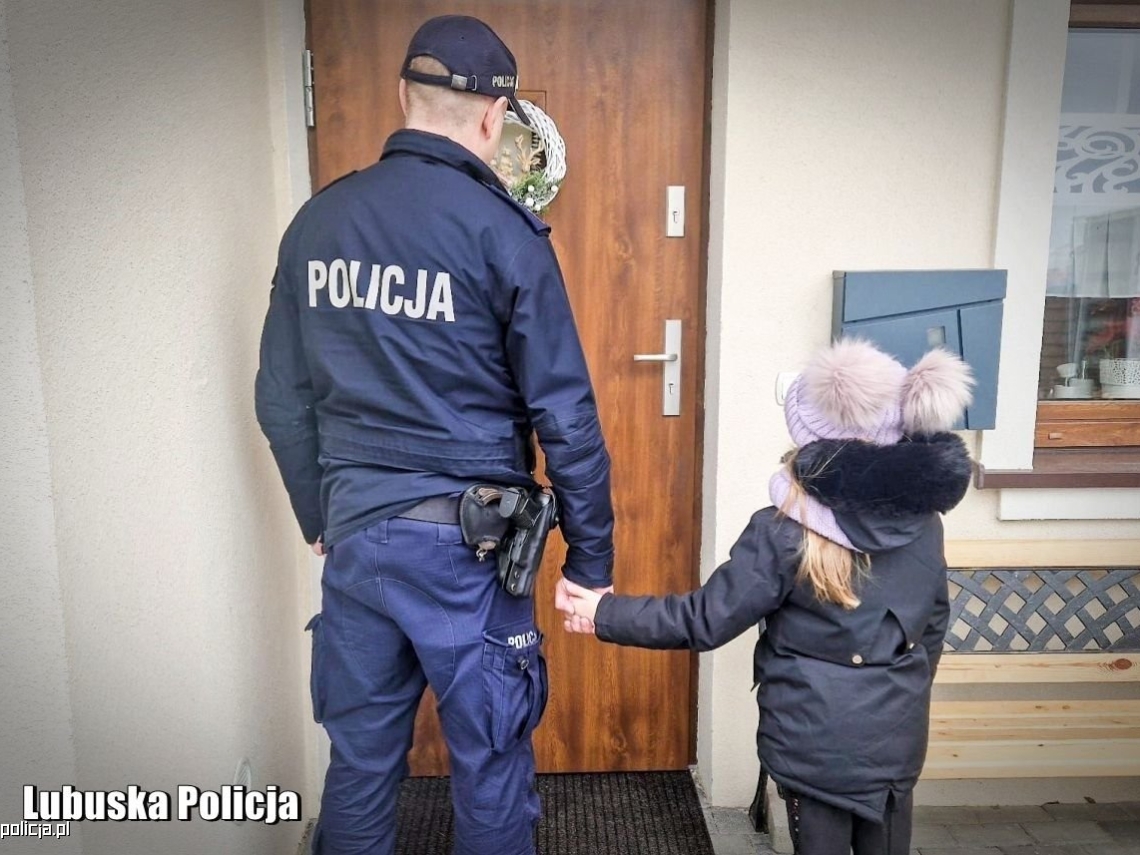 Umundurowany policjant trzyma za rękę dziecko. Oboje stoją przed drzwiami domu.