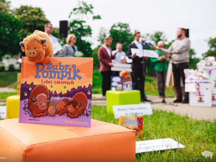 na pierwszym planie książeczka i maskotka żubra - w tle widać osoby podczas konferencji prasowej