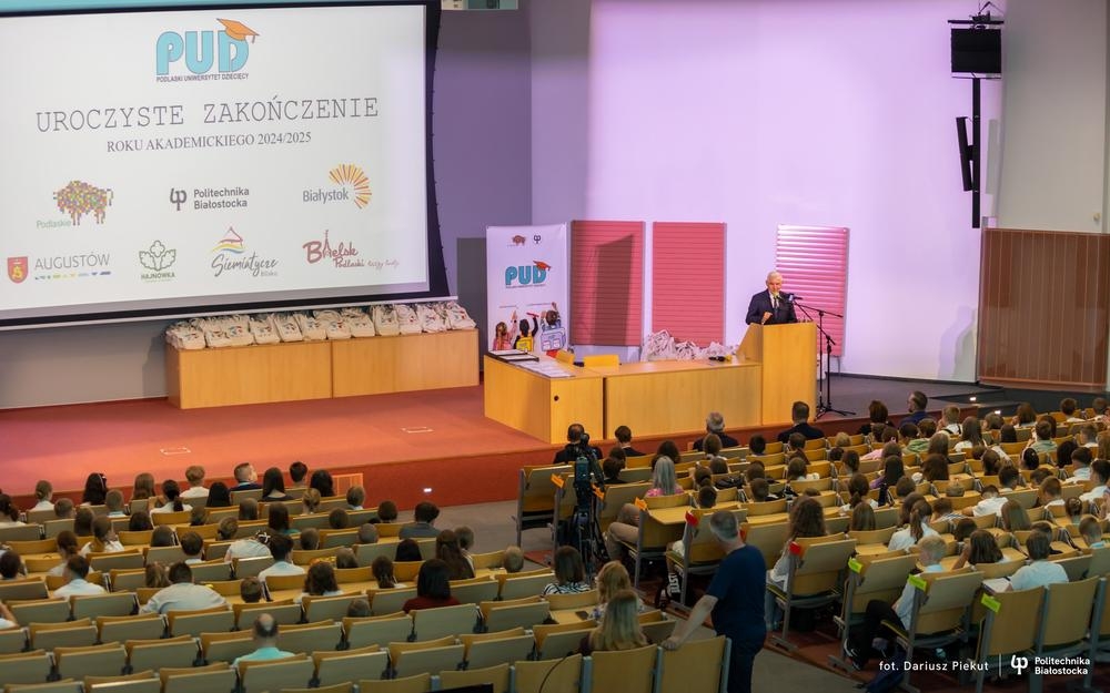 Widok na aulę, w której siedzą dzieci i opiekunowie. Mężczyzna przy mikrofonie.