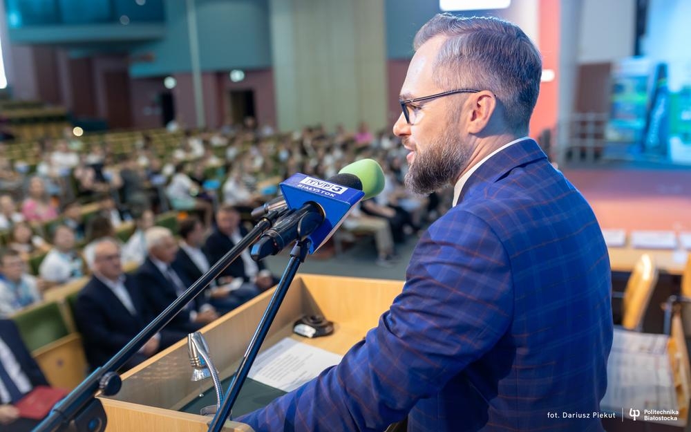 mężczyzna przy mikrofonie. W tle widać osoby siedzące na auli