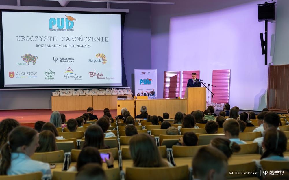 Widok na aulę, w której siedzą dzieci i opiekunowie. Mężczyzna przy mikrofonie.