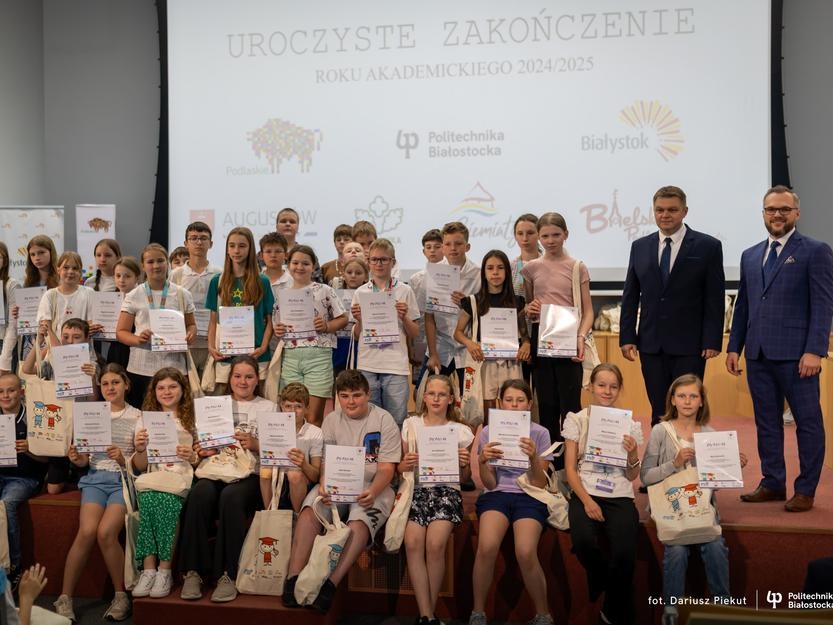 duża grupa dzieci pozuje do wspólnego zdjecia z Burmistrzem Miasta Hajnówka i Prorektorem ds. Studenckich Politechniki Białostockiej. Dzieci trzymają dyplomy zakończenia roku akademickiego.