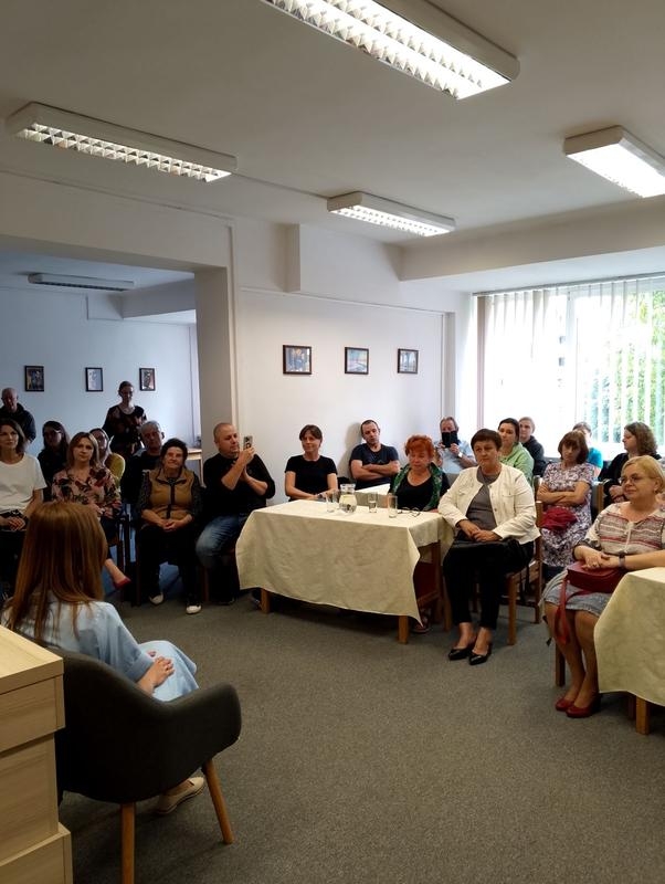 kobieta siedzi przy stoliku, w tle widać słuchaczy i półki z ksiązkami