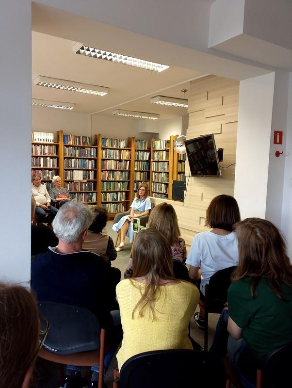 kobieta siedzi przy stoliku, w tle widać słuchaczy i półki z ksiązkami