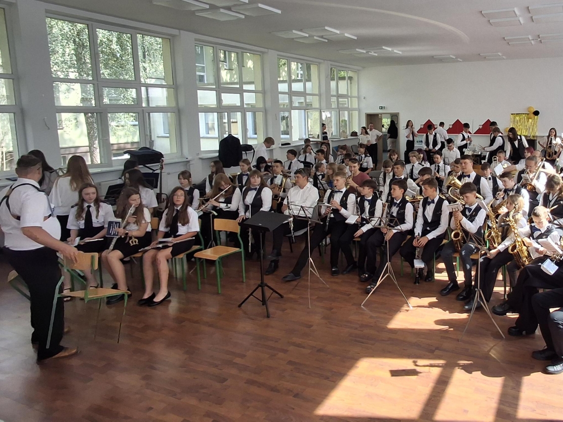 członkowie orkiestry grają na instrumentach. Przed orkiestrą stoi dyrygent