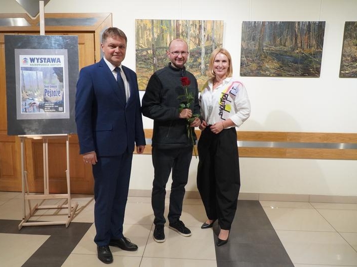 Autor wystawy, burmistrz i jego zastepca pozują do zdjecia. W tle wystawa.