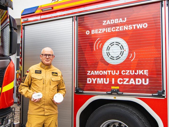 mężczyzna w mundurze stoi przy banerze promującym bezpieczeństwo i pokazuje czujki