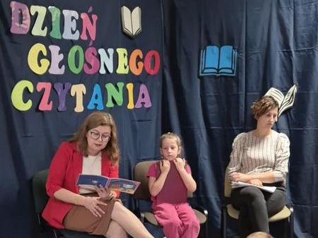 trzy osoby siedzą przy ściance z napisem DZIEŃ GŁOŚNEGO CZYTANIA. Pani Dyrektor czyta z książki.