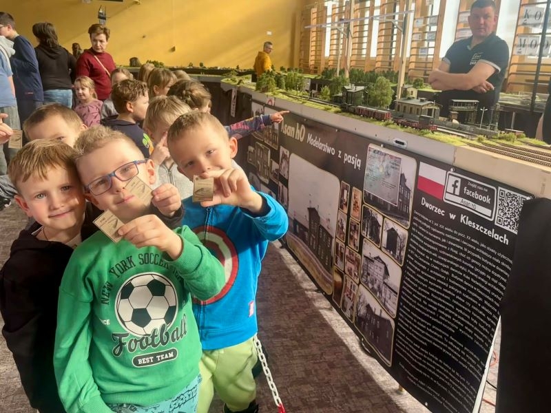 Dzieci oglądają dużą makietę kolejową z wieloma torami, pociągami, lokomotywami, samochodami na drogach, zielenią i miniaturkami budynków.