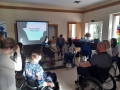 sala szkoleniowa, część uczestników szkolenia siedzi na wózkach inwalidzkich, część stoi obok nich; przed nimi na wózku inwalidzkim stoi prowadząca kurs, za nią stoi ekran z wyświetlonym slajdem