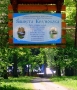 tablica informacyjna zamieszczona nad wejsciem na teren uroczyska; to niebieska tablica zawierająca informację o początka tego miejsca; tuż za nią rozpościera sie zieleń drzew i roślinności