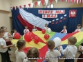 Dzieci z grupy III Puchatki ustawione w półkolu, po lewej stronie dziewczynki w biało - czerwonych spódniczkach w środku chłopczyk trzymający flagę, a po lewej stronie chłopcy. Wszystkie dzieci stoją na baczność śpiewając uroczyście Hymn Polski.