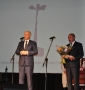 burmistrz wypowiada się do mikrofonu, obok niego stoi laureat trzu mający w rękach bukiet kwiatów