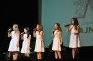 pięć dziewczynek w białych sukienkach wykonuje na scenie piosenkę