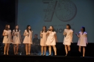 7 dziewczyn w białych sukienkach na scenie wykonują piosenkę
