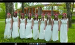 9 dziewczyn w białych długich sukienkach z różowanymi wiankami na głowie, w otoczeniu zieleni traw i drzew