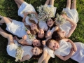 kadr z nagrania teledysku -uśmiechnięte dziewczynki ubrane w białe sukienki z kkwiatami w rękach leża na trawie. 
