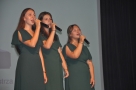 Trzy dziewczyny w zielonych sukienkach śpiewają do mikrofonów. 