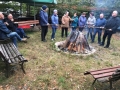 Uczestnicy spotkania stoją przy ognisku i pieką kiełbaski.