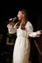 Dziewczyna w białej sukience śpiewa do mikrofonu, który trzyma w ręku.