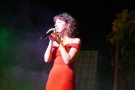 Dziewczyna w czerwonej sukience śpiewa do mikrofonu, który trzyma w ręku.