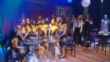 Dziewczęta w zółtych bluzkach stoją przy stolikach. Sceneria jak w kawiarni.