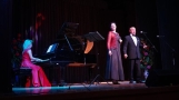 Kobieta i meżczyzną stoją przed fortepianem, na którym gra kobieta. Wszyscy ubrani w stroje wieczorowe.