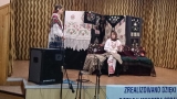 dwie osoby w stojach ludowych na scenie. W tle widać zawieszone haftowane makaty i chusty