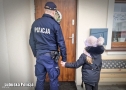 Umundurowany policjant trzyma za rękę dziecko. Oboje stoją przed drzwiami domu.