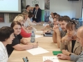 Przy stole siedzi grupa osób. W tle widać burmistrza rozmawiającego z prowadzącą spotkanie.