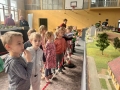 Dzieci oglądają dużą makietę kolejową z wieloma torami, pociągami, lokomotywami, samochodami na drogach, zielenią i miniaturkami budynków.