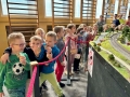 Dzieci oglądają dużą makietę kolejową z wieloma torami, pociągami, lokomotywami, samochodami na drogach, zielenią i miniaturkami budynków.