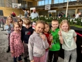 Dzieci oglądają dużą makietę kolejową z wieloma torami, pociągami, lokomotywami, samochodami na drogach, zielenią i miniaturkami budynków.
