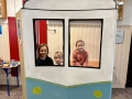  dzieci są w foto budce w kształcie lokomotywy. Wyglądają przez okienko i uśmiechają się do zdjęć.