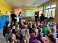 dzieci siedzą na ławkach w szkolnym korytarzu, oglądają film edukacyjny oraz pokaz psa patrolowego.