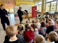 dzieci siedzą na ławkach w szkolnym korytarzu, oglądają film edukacyjny oraz pokaz psa patrolowego.